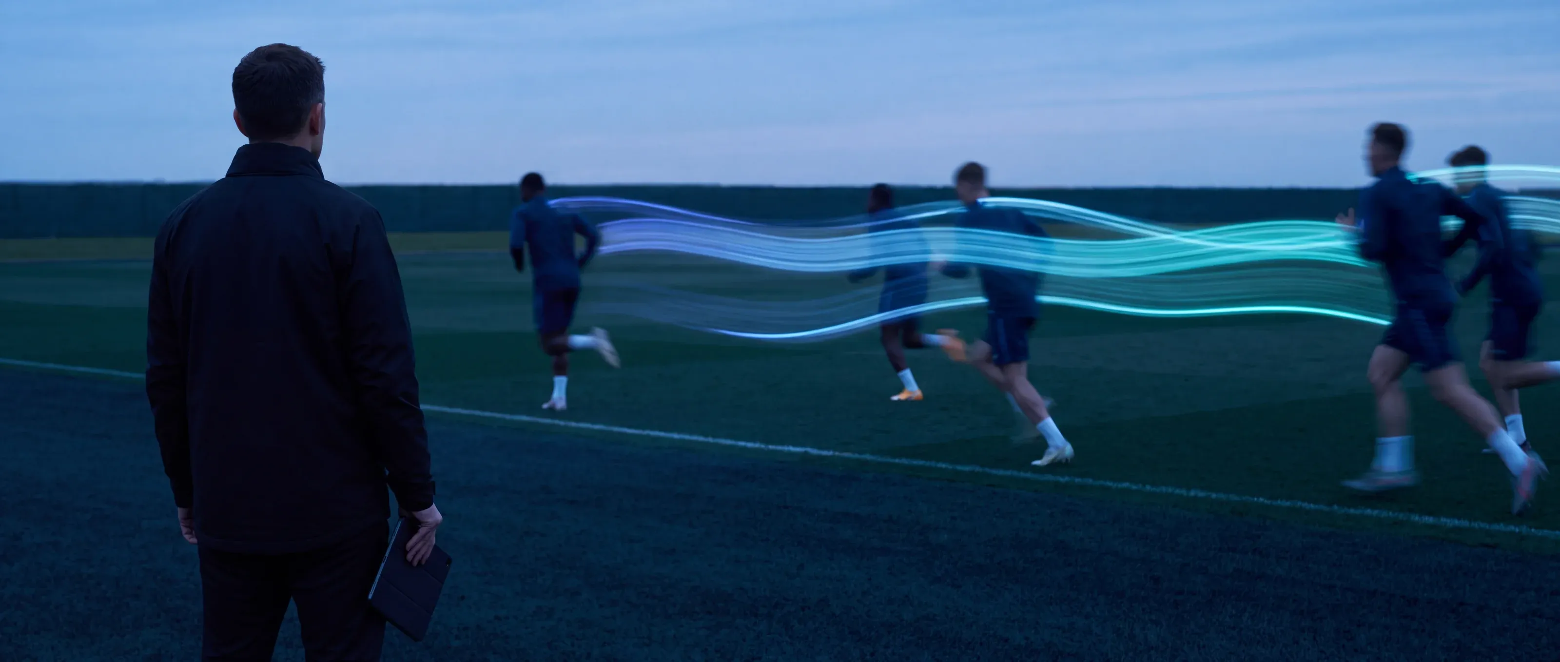 A coach stands at the side of a training pitch at dusk, seen from behind, with a tablet in one hand. Athletes run past, and each leaves a line of blue light behind.